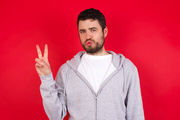 young handsome caucasian man in sports clothes against red background makes peace gesture keeps lips folded shows v sign. Body language concept