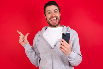 Astonished young handsome caucasian man in sports clothes against red background holding her telephone and pointing with finger aside at empty copy space