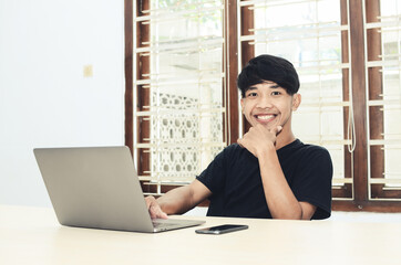 The Asian man who is sitting at the office table wearing a black shirt is smiling in front of the laptop