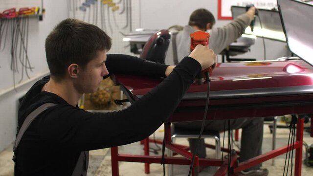 Work in car repair shot of two man's - PDR technology professionals using special tools and methods removing dents use no painting. Younger guy using an industrial dryer heating a red car door surface