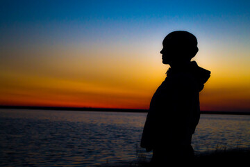 Fototapeta premium The girl at sunset stands at the shore and looks into the distance, waiting for a miracle.