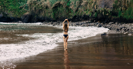 Fit woman in bikini with her back  walking on black beach. Wild nature, empty bay. 