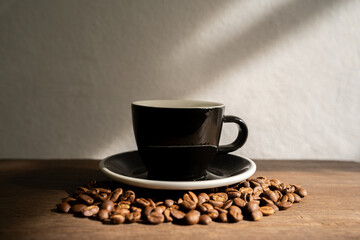 Black cup on a wooden table, Beautiful light and shadow on the white wall, Many coffee beans placed all around, Morning coffee break, Selective focus, Latte, Cup, Horizontal orientation, Copy room.