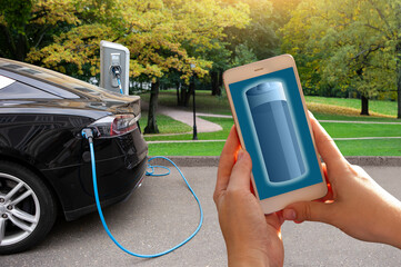 Hands with smartphone on a background of a car at the charging station for electric vehicles. Battery level on the phone screen