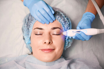 Beautician hands using aqua peel water galvanic device. Female client in disposable shower hat...
