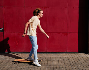 a young man with curly hair using his skateboard with a red background behind him on a sunny day