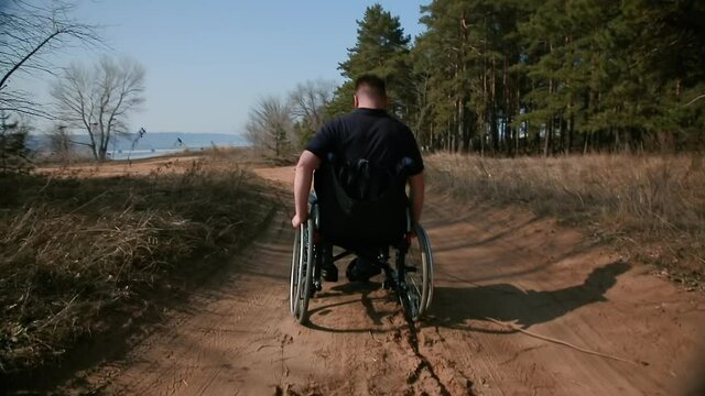 A man rides in a wheelchair on the road. A disabled man in a wheelchair rides heavily on a sandy road along the forest. Rear view. Selective focus