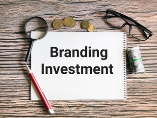 Business and finance concept. Selective focus notebook written branding investment with pen,coins,fake money,magnifying glass and eye glasses.