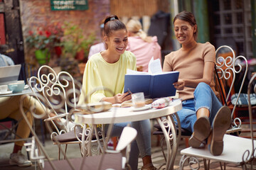 Two female students enjoying an interesting book while have a drink in bar's garden. Leisure, bar, friendship, outdoor