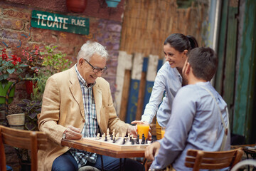 An elderly man is served by beautiful waitress while he plays a chess with his young friend in the bar. Leisure, bar, friendship, outdoor