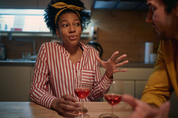 young afro-american woman with scarf on her head, talking with caucasian man, explaining, drinking red win. Friendship at home during coronavirus lockdown