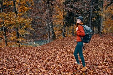 happy young woman with a backpack in jeans boots and a sweater are walking in the autumn forest near the tall trees 