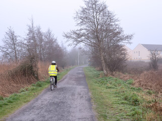 Woman in high visibility vest and helmet riding bicycle on a small road into fog. Safety on the road concept. Outdoor sport and healthy life style