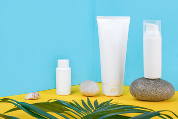 White blank cosmetic tube, bottle with sunscreen, sun cream, moisturizing lotion, rock and green leaf on blue, yellow background. Concept skin care in sea beach holidays in summer vacation