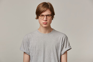 Teenage guy, upset looking man with blond hair. Wearing grey t-shirt and glasses. People and emotion concept. Frowns and pouts his lip. Watching at the camera isolated over grey background
