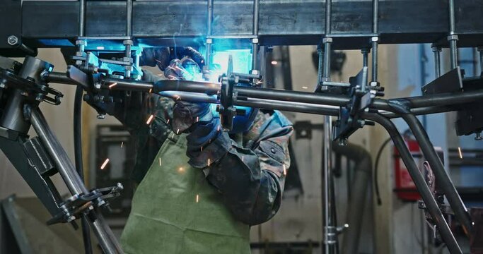 Male mechanic welding metal structure. Man in apron lowering protective mas and welding metal structure during work in garage