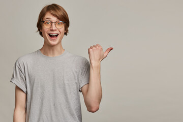 Teenage guy, happy looking man with blond hair. Wearing grey t-shirt, glasses and has braces. Watching at the camera and pointing with thumb to the right at copy space, isolated over grey background
