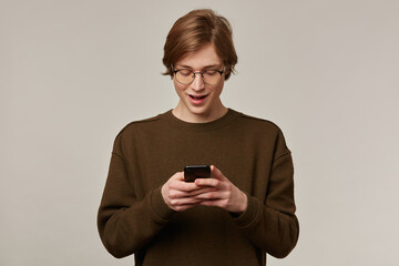 Teenage guy, happy looking man with blond hair. Wearing brown sweater and glasses. Holding a smartphone and looking at it. Typing a message. Stand isolated over grey background