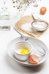 Eggshell, yolk and whisk in ceramic bowl. Flour and sieve on paper. Egg and water bottle.