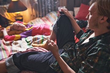 Fototapeta na wymiar Young man smoking a cigarette and drinking wine sitting on the bed with his girlfriend relaxing near by him at home