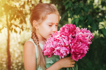 Fototapeta premium portrait of a happy cute little caucasian seven year old kid girl, holds in hands and smell and enjoying a bouquet of pink peony flowers in full bloom on the background of nature. flare