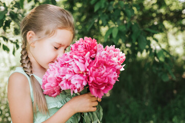 Fototapeta premium portrait of a happy cute little caucasian seven year old kid girl, holds in hands and smell and enjoying a bouquet of pink peony flowers in full bloom on the background of nature