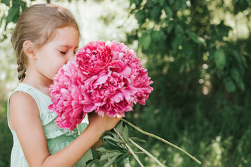 portrait of a happy cute little caucasian seven year old kid girl, holds in hands and smell and enjoying a bouquet of pink peony flowers in full bloom on the background of nature