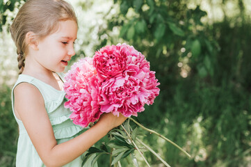 portrait of a happy cute little caucasian seven year old kid girl, holds in hands and smell and enjoying a bouquet of pink peony flowers in full bloom on the background of nature