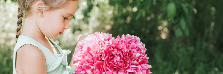 portrait of a happy cute little caucasian seven year old kid girl, holds in hands and love to see a bouquet of pink peony flowers in full bloom on the background of nature. banner
