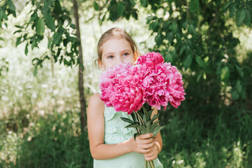 portrait of a happy cute little caucasian seven year old kid girl in light green sundress, holds in hands a bouquet of pink peony flowers in full bloom on the background of nature