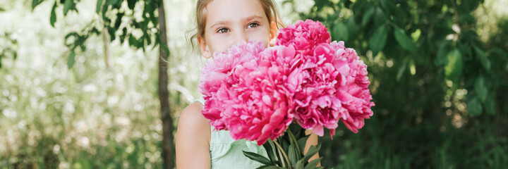 portrait of a happy cute little caucasian seven year old kid girl in light green sundress, holds in hands a bouquet of pink peony flowers in full bloom on the background of nature. banner