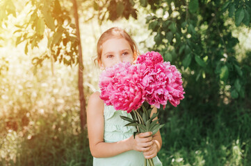 portrait of a happy cute little caucasian seven year old kid girl in light green sundress, holds in hands a bouquet of pink peony flowers in full bloom on the background of nature. flare