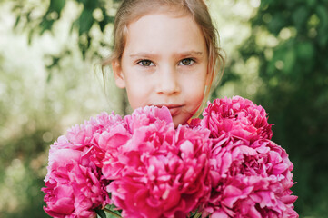 Fototapeta premium portrait of a happy cute little caucasian seven year old kid girl, holds in hands a bouquet of pink peony flowers in full bloom on the green background of nature