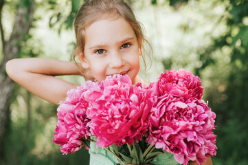 Fototapeta premium portrait of a happy cute little caucasian seven year old kid girl, holds in hands a bouquet of pink peony flowers in full bloom and straightening her hair on the background of nature