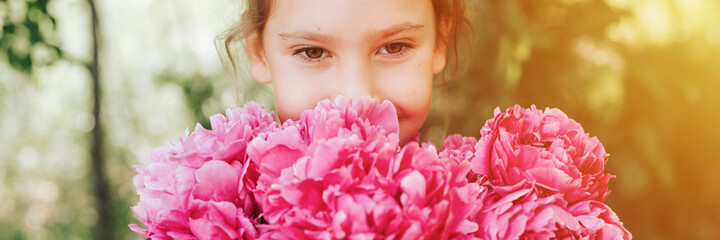 Fototapeta premium portrait of a happy cute little caucasian seven year old kid girl, holds in hands and smell and enjoying a bouquet of pink peony flowers in full bloom on the background of nature. banner. flare