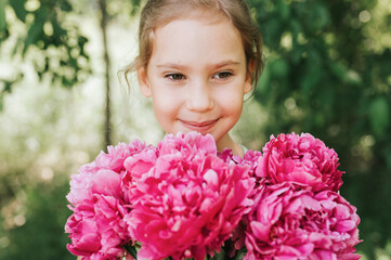 Fototapeta premium portrait of a happy cute little caucasian seven year old kid girl, holds in hands a bouquet of pink peony flowers in full bloom on the green background of nature