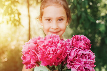 Fototapeta premium portrait of a happy cute little caucasian seven year old kid girl, holds in hands a bouquet of pink peony flowers in full bloom on the green background of nature