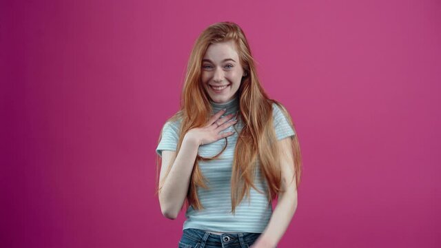 The red-haired, amused young woman smiles broadly at a good joke, pointing her finger at the source of this joke, in a blue casual T-shirt, isolated on a pink background. The concept of people's