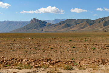 View of a stone desert field in front of beautiful brown vegetation-free rolling mountains and blue sky with little white clouds between Midelt and the Gorges du Ziz in Morocco, Africa
