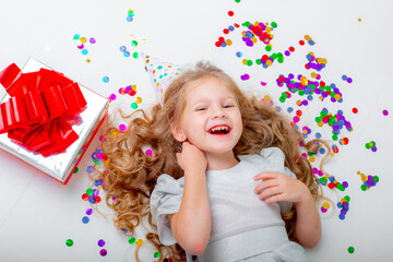 happy little girl in a birthday cap lies on back with gifts and confetti on the floor on a white background.top view