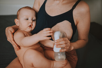 Young mother with newborn cute infant naked baby boy, holding him on arms, using breast pump hugging and breastfeeds with breast milk. Healthy child, concept of hospital and happy motherhood. Nursery