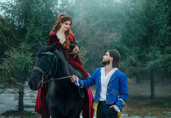 Fototapeta na wymiar Medieval couple in love. man meets woman and gives rose in winter forest. Vintage clothing red long dress. Blue costume tailcoat caftan. Prince and princess together. Black horse. redhead hairstyle.