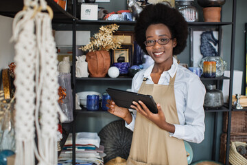 Close up of smiling pretty afro american woman in eyegasses using digital tablet for making inventory at decor shop. Attractive saleswoman in apron standing with modern gadget in hands. - obrazy, fototapety, plakaty