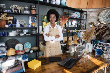 Fototapeta na wymiar Portrait of likable afro american woman in uniform and eyeglasses smiling and looking at camera. Modern shop with various decor on shelves.