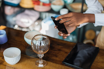 Close up of black saleswoman using modern smartphone for taking photo of shiny glass at store. Worker making website with available assortment of decor shop.