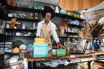 Qualified afro american saleswoman in eyeglasses packing boxes with various souvenirs for clients....
