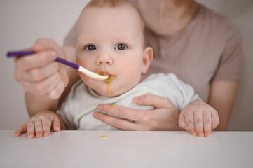 Little newborn funny baby boy learning to eat vegetable or fruit puree from glass jar with spoon. Young mother helping little son eat first food