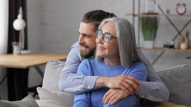 Handsome smiling adult man hugging beautiful senior mature mother, looking at window. Happiness, love and care.Two generations family close up portrait. Mothers day concept.