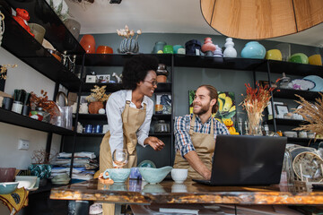 Smiling african woman and caucasian man working together st decor shop and using laptop. Two colleagues doing inventory of goods with help of modern gadget.