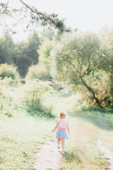 little girl 2-3 years old European appearance. the child walks on a path near the forest. blonde hair. green forest and grass. sunny day.